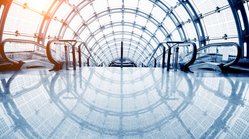 This brightly lit photo shows the the reflective surface of a floor headed toward the top of a stairway descending into a tunnel covered by glass